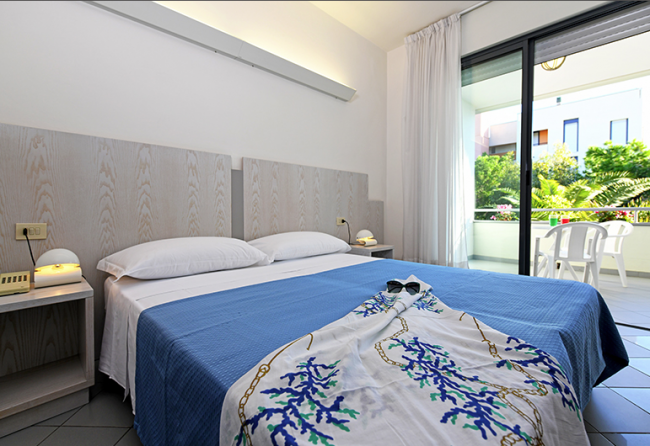 Camera d'albergo moderna con letto matrimoniale, copriletto blu e balcone con vista su un giardino.