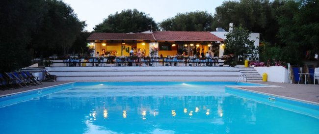 Piscina illuminata di fronte a un edificio con veranda e ospiti seduti.