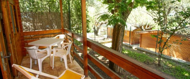 Veranda esterna in legno con tavolo e sedie bianche, immersa nel verde.