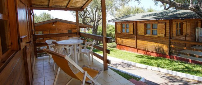Veranda con tavolo e sedie di plastica affacciata su un giardino con bungalow in legno.