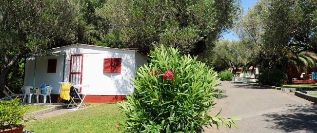 Bungalow bianco con infissi rossi circondato da alberi e piante fiorite.