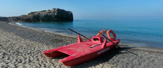 Spiaggia con pedalò rosso sulla sabbia. Sfondo di mare calmo e scoglio.