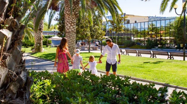 Una famiglia cammina in un parco soleggiato con palme, priva di persone intorno.