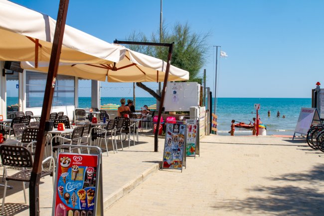 Bar sulla spiaggia con sedie sotto tende da sole, vicino al mare con bagnanti.