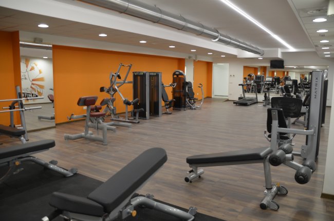 Palestra moderna con attrezzi e pesi su pavimento in legno, pareti arancioni e specchi.