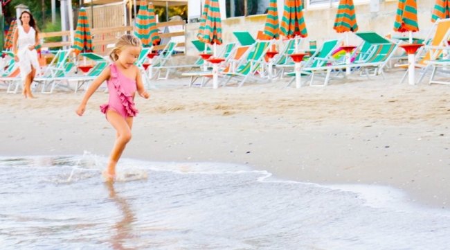 Bambina corre sorridendo verso l'acqua sulla spiaggia, mentre una donna la osserva in lontananza.