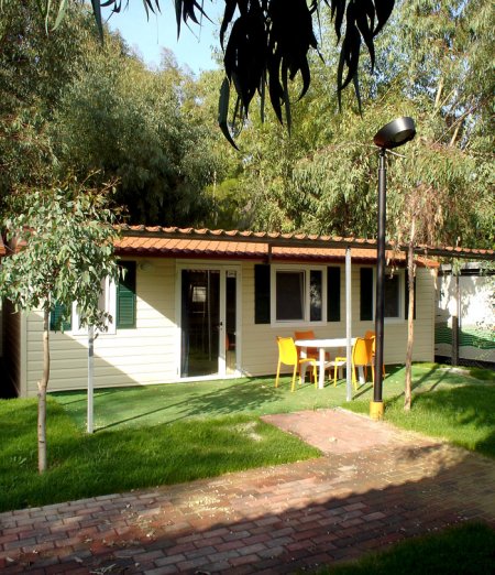 Una casetta con terrazza, sedie arancioni e tavolo su prato verde circondata da alberi.