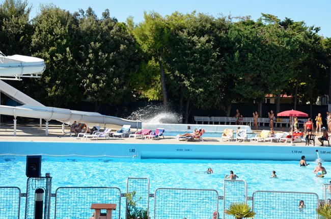 Grande piscina con scivoli d'acqua, persone che nuotano e si rilassano su lettini.