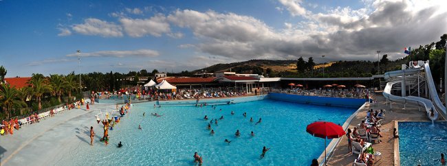 Piscina affollata con scivoli e palme, cielo parzialmente nuvoloso.