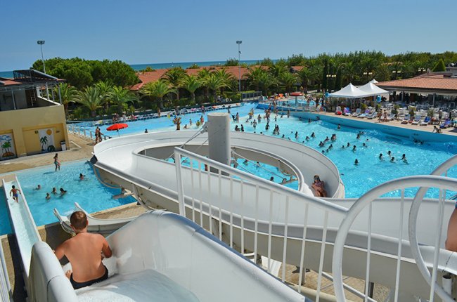 Persona su scivolo d'acqua, vista di piscina affollata e palme.