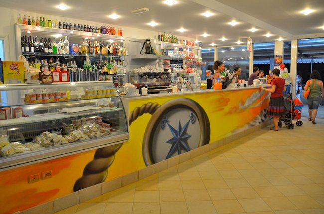 Interno di un bar luminoso, con un bancone colorato e persone che ordinano.