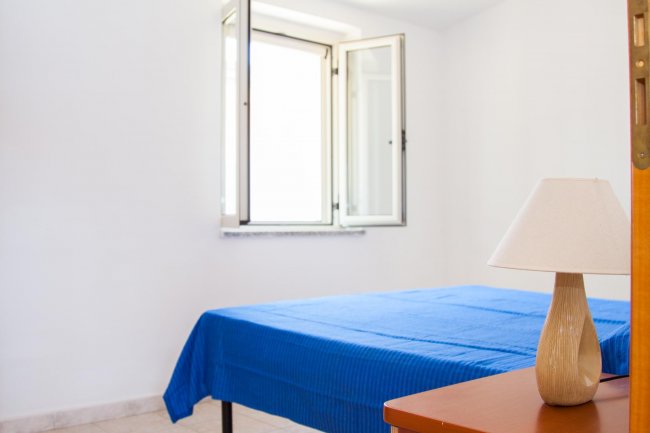 Camera semplice con letto blu, finestra aperta e lampada su un comodino.
