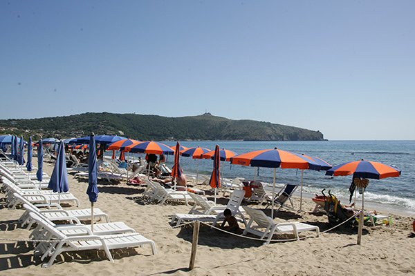 Spiaggia sabbiosa con lettini bianchi e ombrelloni blu e arancioni, vista su collina.