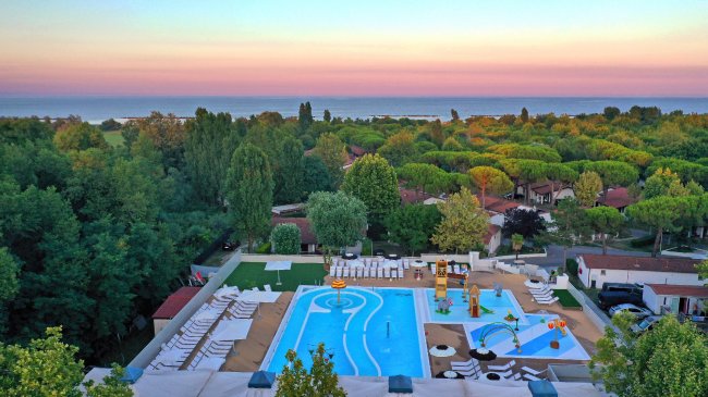 Vista dall'alto di una piscina e spa con lettini, circondata da alberi e un panorama al tramonto.
