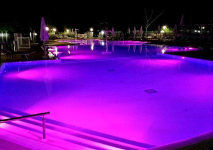 Piscina illuminata di notte con luci viola e sedie a sdraio circostanti.