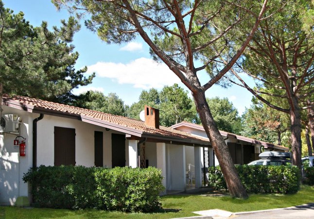 Bungalow bianchi con tetti rossi, circondati da alberi e piante verdi, sotto un cielo sereno.
