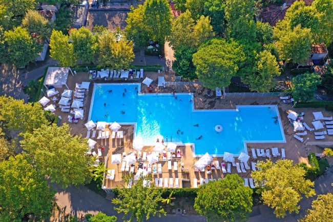 Piscina grande a forma irregolare circondata da ombrelloni e lettini, immersa nel verde.