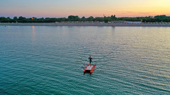 Persona su pedalò rosso in mare al tramonto con spiaggia sullo sfondo.