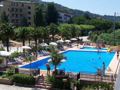 Piscina all'aperto circondata da sdraio e ombrelloni, in un'area verde con edifici vicini.
