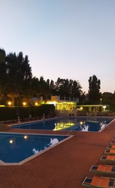 Piscina al tramonto con luci accese e sdraio arancioni.