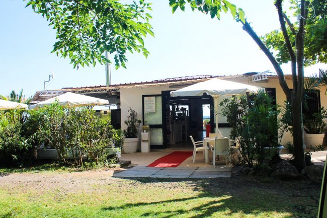 Ristorante con ombrelloni, tavolini e giardino con piante e alberi.