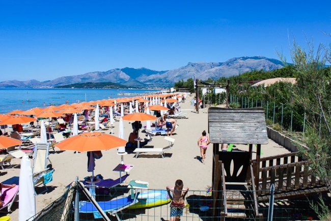 Spiaggia con ombrelloni arancioni, persone su lettini e monti sullo sfondo.