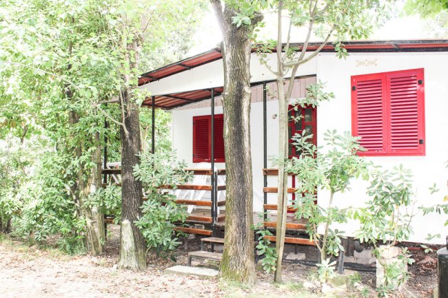 Piccola casa bianca con finestre rosse e veranda, circondata da alberi verdi.