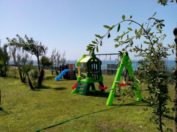 Area giochi per bambini su erba verde, con scivoli e altalene. Mare e alberi sullo sfondo.