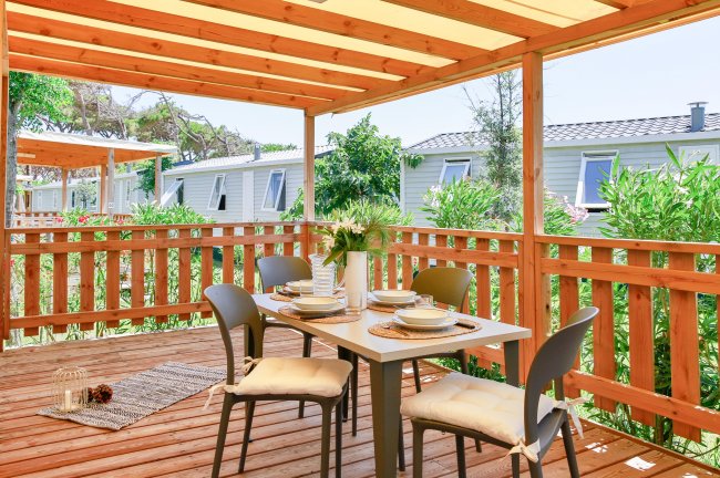 Un patio in legno con tavolo apparecchiato e sedie, circondato da vegetazione.