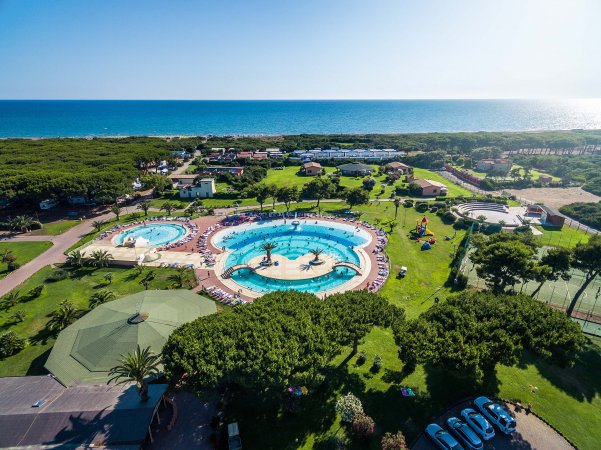 Vista dall'alto di un complesso con piscine circolari, prato, e mare sullo sfondo.
