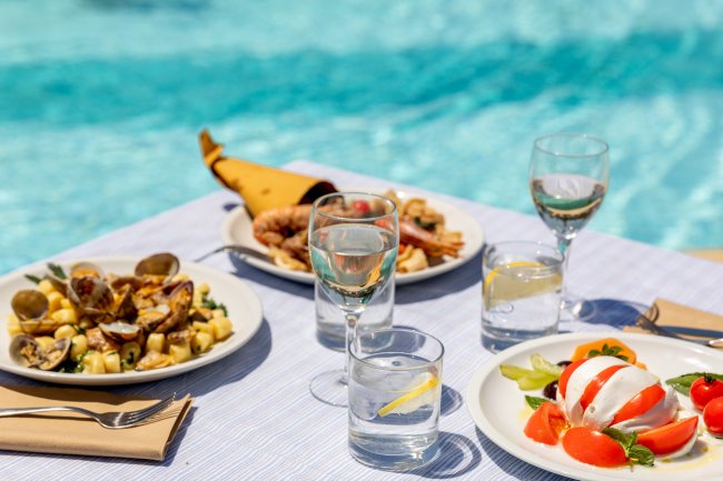 Piatti di cibo con frutti di mare e caprese, bicchieri di vino e acqua, davanti a una piscina.