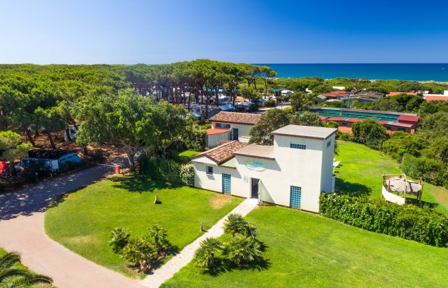 Edificio bianco su un prato verde, circondato da alberi e strutture, con vista sul mare.