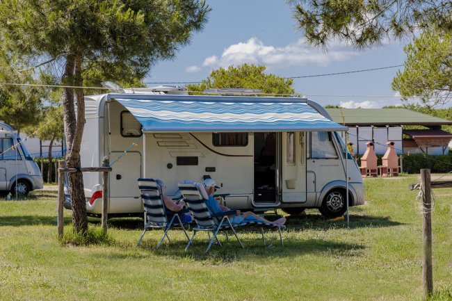 Camper parcheggiato su prato, sotto una tenda blu a strisce, con persone sedute su sedie a sdraio.