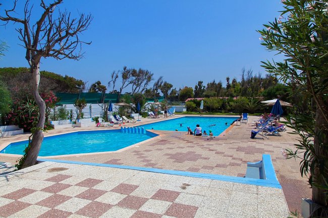 Due piscine all'aperto con lettini e ombrelloni, circondate da vegetazione e un cielo blu.