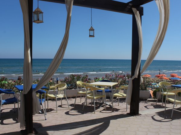 Tavoli esterni con vista mare, tende e lanterne, spiaggia con ombrelloni sullo sfondo.