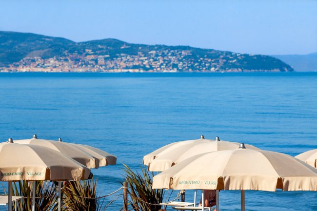 Ombrelloni beige su una spiaggia con vista sul mare e un promontorio in lontananza.