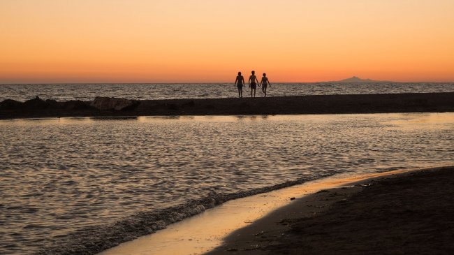Tre persone camminano su una spiaggia al tramonto, con il mare sullo sfondo.