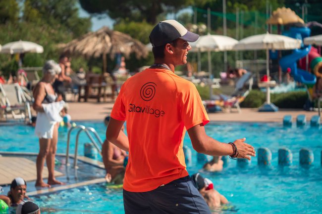 Persona con maglietta arancione in piscina, diverse persone nuotano.