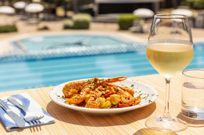 Tavolo con piatto di gamberi e pasta, bicchiere di vino bianco vicino a una piscina.