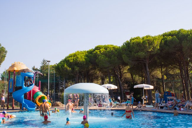 Piscina con giochi d'acqua e scivolo colorato, persone che nuotano.