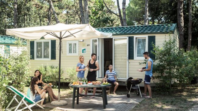 Gruppo di amici fuori da una roulotte, intorno a un tavolo all'aperto, con barbecue e ombrellone.