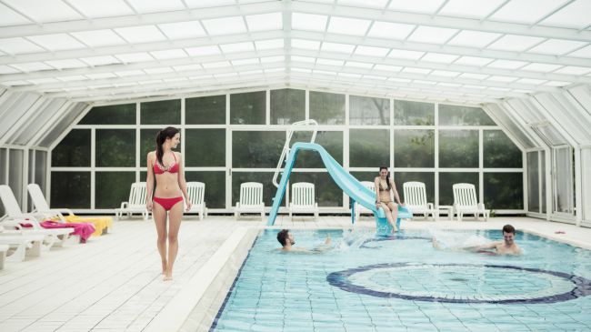 Interno di una piscina coperta con persone che nuotano e una donna sul bordo.