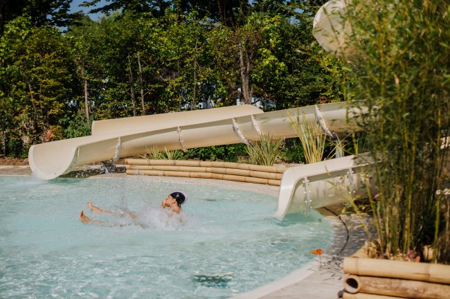 Un bambino si tuffa in piscina uscendo dallo scivolo, circondato da vegetazione.