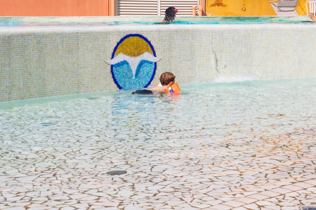 Bambino con braccioli arancioni in una piscina, accanto a un bordo con un logo a mosaico.