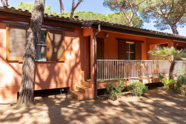 Bungalow arancione con terrazza, finestre con persiane in legno e vegetazione circostante.