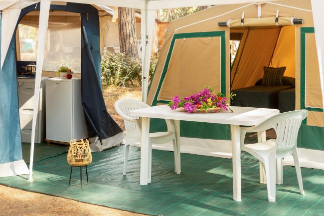 Esterno di una tenda da campeggio con tavolo e sedie bianche, fiori su tavolo, mini frigo in vista.