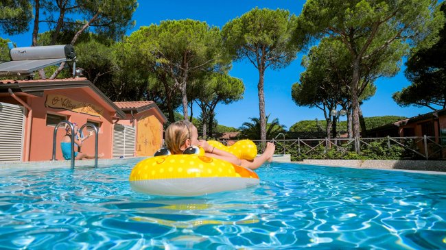 Una bambina su un gonfiabile giallo in una piscina, circondata da pini.