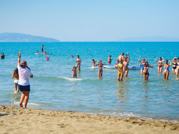 Spiaggia affollata con persone che giocano in acqua limpida. Mare azzurro e cielo sereno.