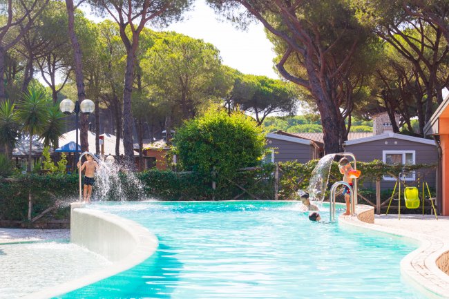 Bambini giocano in una piscina con giochi d'acqua e alberi intorno.