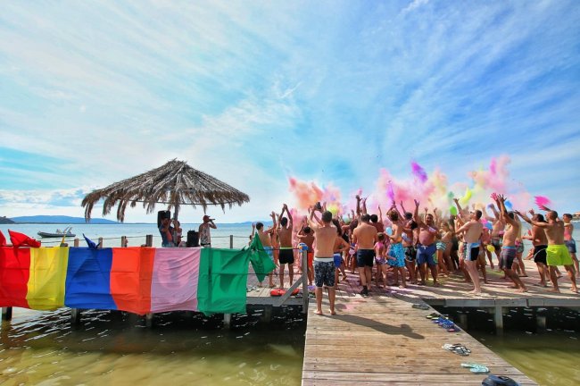 Persone su un molo festeggiano lanciando polveri colorate, sotto un cielo azzurro.